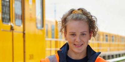 Auszubildende Ronja in BVG-Arbeitskleidung und Warnweste steht vor einer U-Bahn neben den Gleisen