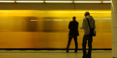 Zwei Männer stehen in einem U-Bahnhof. Eine Bahn fährt gerade ein und ist verschwommen im Hintergrund zu sehen.