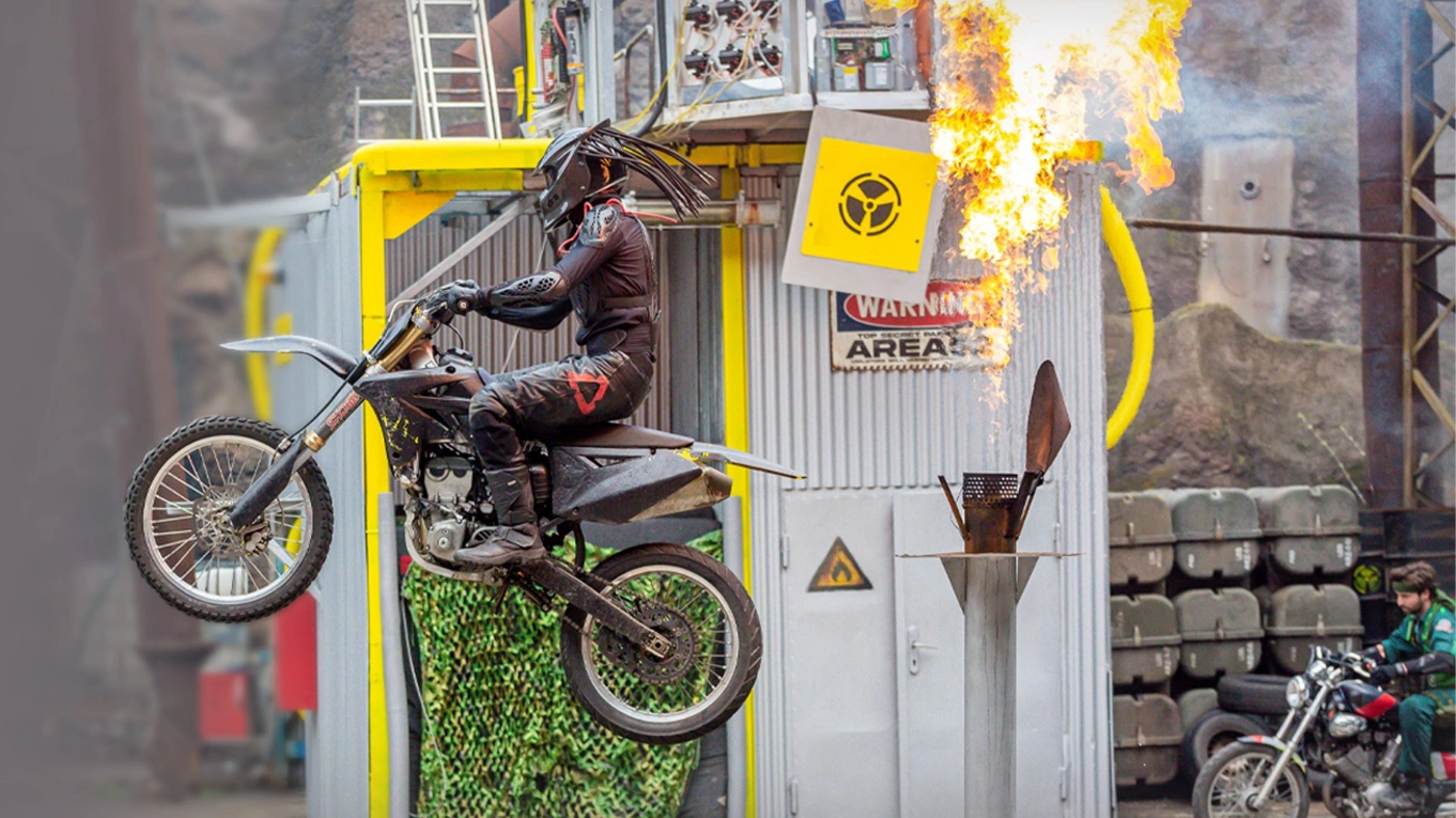 Eine Aufnahme eines Motorrad-Stunts im Filmpark Babelsberg. Ein in schwarz gekleideter Motorradfahrer fliegt durch die Luft, im Hintergrund schießen Flammen in die Höhe.