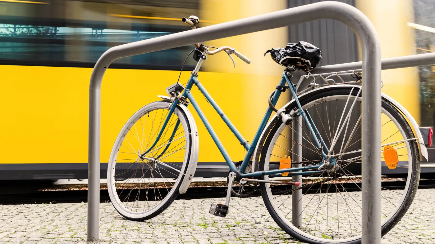 Ein Fahrrad steht an einem Fahrradständer einer Haltestelle. Im Hintergrund fährt eine Tram. 