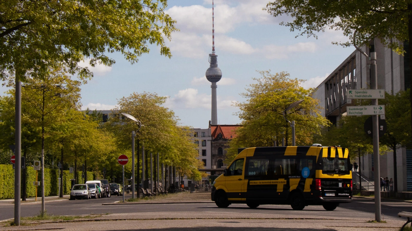 Blick auf eine Berliner Straße, auf der gerade ein BVG Muva fährt. Im Hintergund thront der Berliner Fernsehturm.